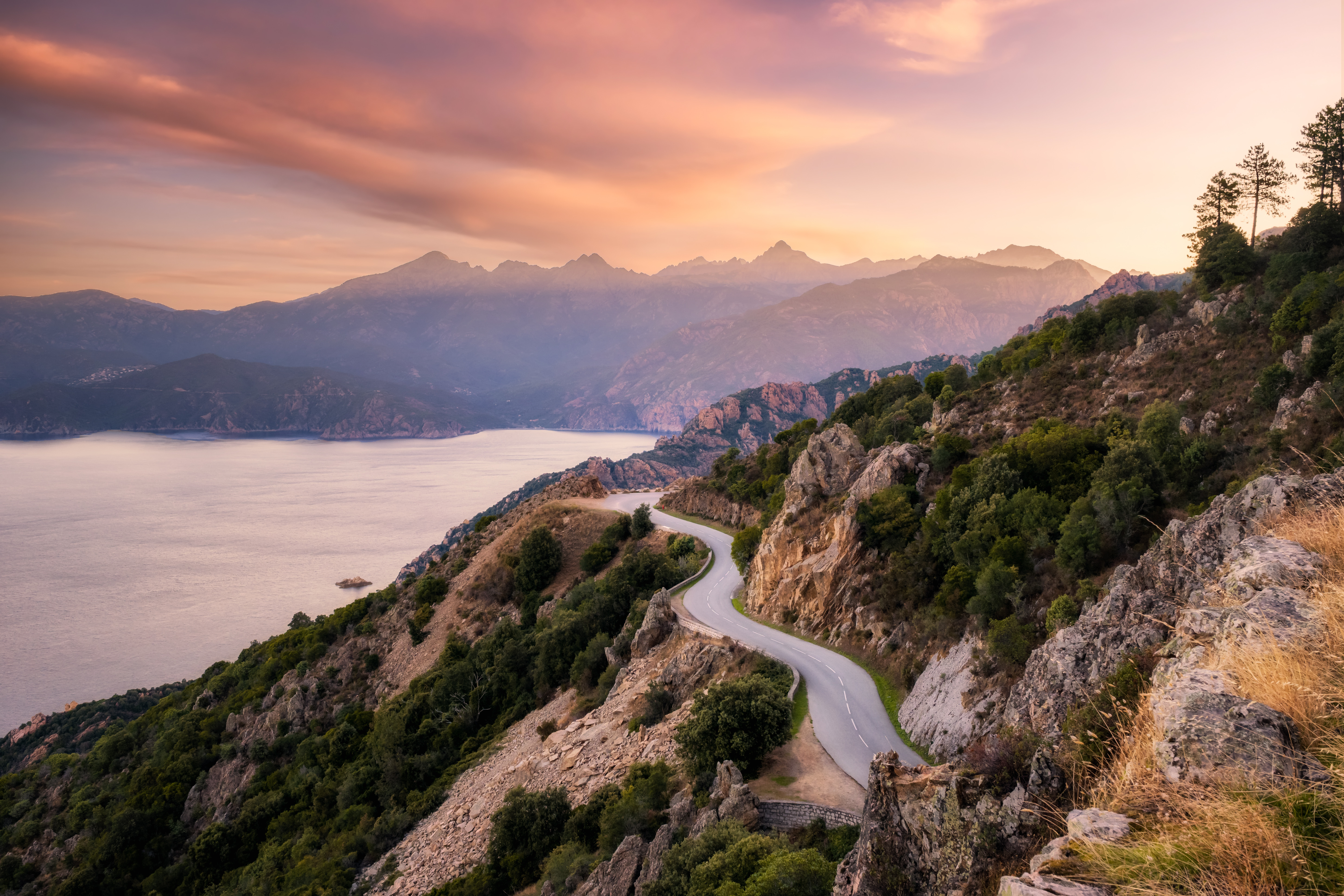 The D824 road winding its way along the coast from Capu Rossu towards Piana on the west coast of Corsica as the early morning sun lights up the distant mountains